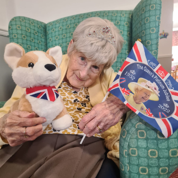 Quiz Queen Dora of Pennington Court wearing a tiara, with a Corgi stuffed toy and holding a small Union Jack flag with the face of Queen Elizabeth on it.